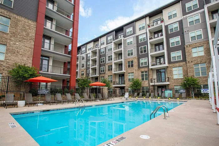 Modern apartment complex featuring a sparkling outdoor pool with lounge chairs, vibrant orange umbrellas, and elegant balconies with scenic views.