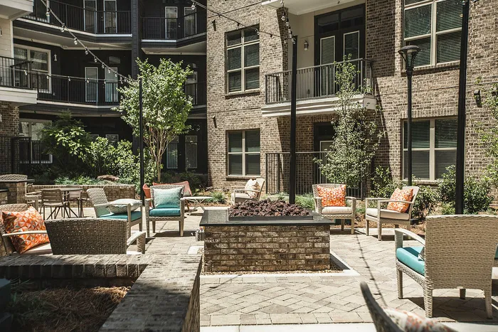 Charming outdoor courtyard with cozy wicker seating, vibrant cushions, and string lights, surrounded by lush greenery and modern brick architecture.