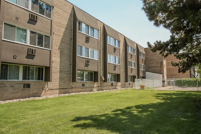 Charming apartment complex with brick exterior and large windows, surrounded by lush greenery and a well-maintained lawn.