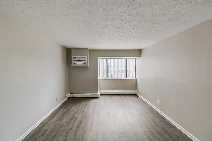 Bright and airy empty living room featuring sleek hardwood floors, neutral walls, large window with vertical blinds, and a wall-mounted air conditioning unit.
