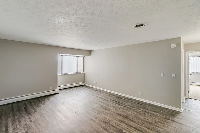 Bright and spacious living area with sleek hardwood floors, large window, and neutral walls, seamlessly leading into a cozy room.