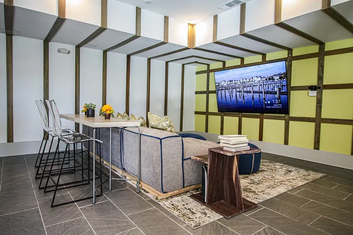 Modern living room with sleek design featuring a large flat-screen TV, chic blue-gray sofa, and a stylish bar table with clear chairs and vibrant decor.