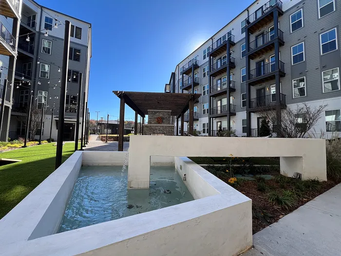 Modern apartment complex courtyard with sleek architecture, water feature, and lush green landscaping under a clear blue sky.