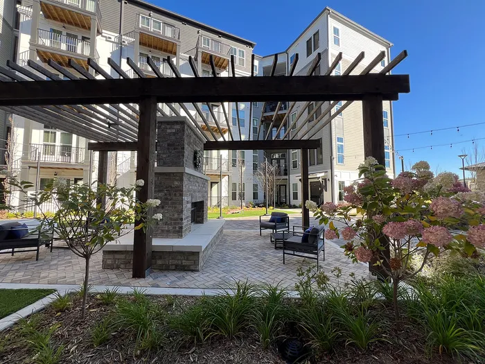 Elegant outdoor courtyard featuring a pergola, modern seating area, and lush landscaping with flowering plants against a backdrop of contemporary apartments.