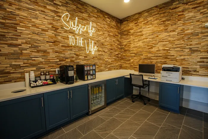 Cozy and stylish community workspace featuring rustic wood accent wall, modern blue cabinetry, coffee station, and office essentials.