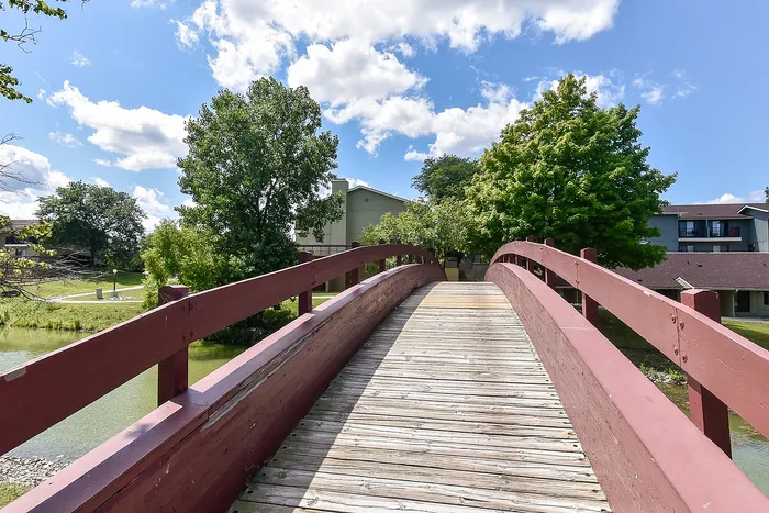 Charming wooden footbridge leading to scenic residential area, surrounded by lush greenery and vibrant blue skies.