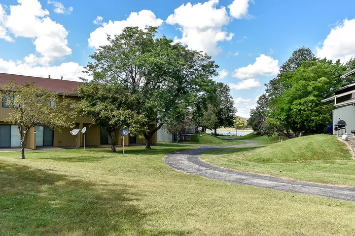 Charming apartment complex with lush greenery, a winding path, and serene surroundings under a bright blue sky.