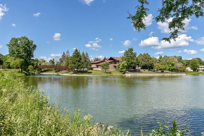 Charming lakeside residence with lush greenery, picturesque views, and a serene wooden bridge under a clear blue sky.