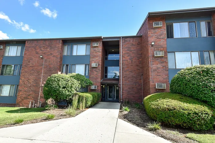 Charming brick apartment building with lush green landscaping and a welcoming entrance under a bright blue sky.