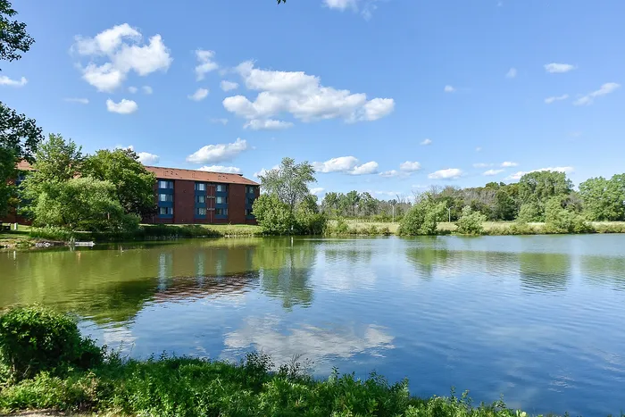 Charming lakeside view with a modern brick apartment building surrounded by lush greenery and blue skies.