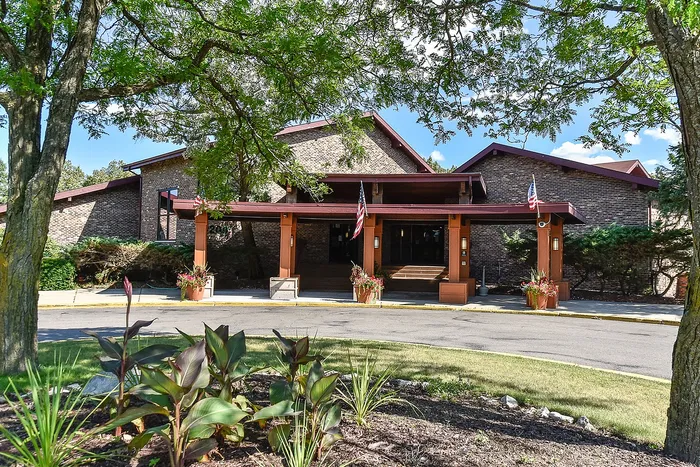 Charming and welcoming brick building entrance with beautiful landscaped gardens and flags flying, set under a clear, blue sky.