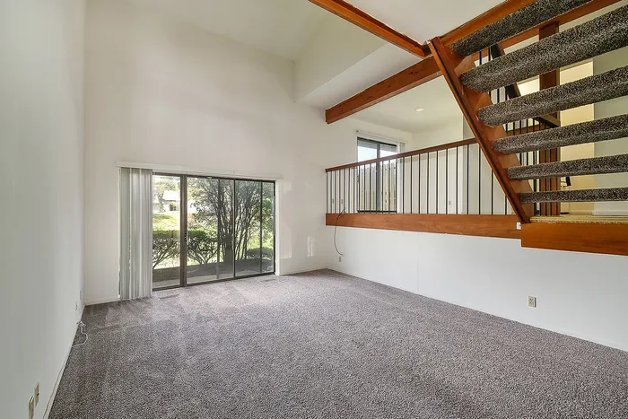 Bright and airy living room with carpeted floors, large sliding glass doors, and a modern mezzanine featuring wooden accents and stylish railings.