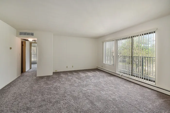 Bright and spacious living room with large windows, plush carpet, and a private balcony overlooking lush greenery.