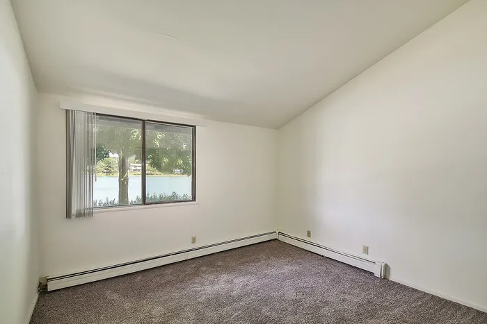 Bright bedroom with a serene lake view through a wide window, featuring soft carpet flooring and neutral walls.