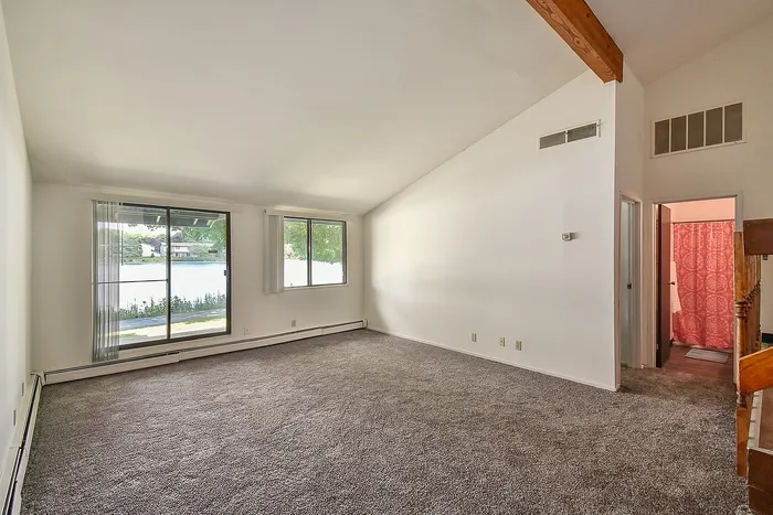 Bright living room with plush carpet, large windows overlooking serene outdoor view, and a stylish wooden beam ceiling accent.