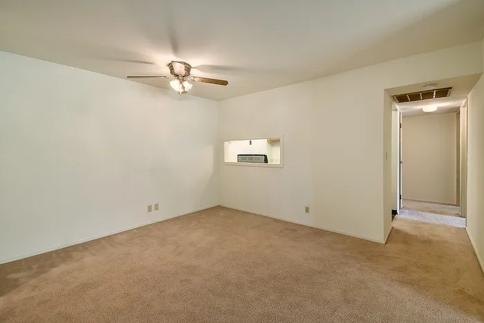 Inviting living area with soft carpet flooring and a ceiling fan, featuring a cozy pass-through window to the kitchen.