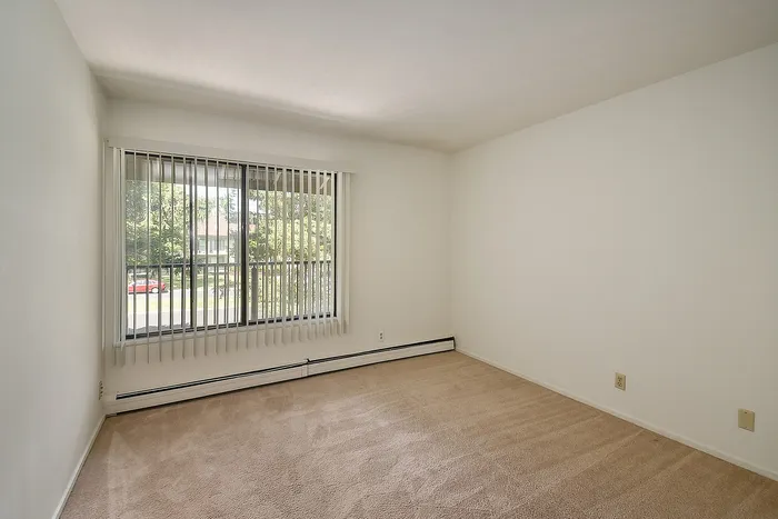 Bright bedroom with large window and vertical blinds, offering a serene view of lush greenery.
