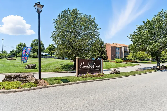 Inviting entrance to Boulder Creek Apartments with lush greenery, welcoming flags, and a sunny blue sky.