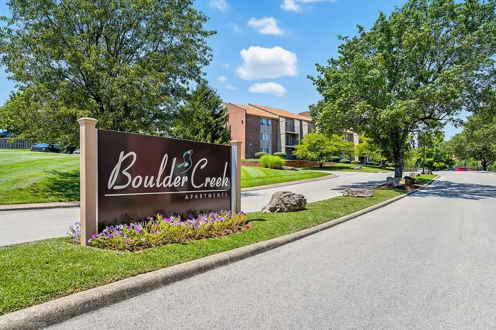 Boulder Creek Apartments entrance sign surrounded by lush greenery and colorful flowers on a sunny day, with modern apartment buildings in the background.