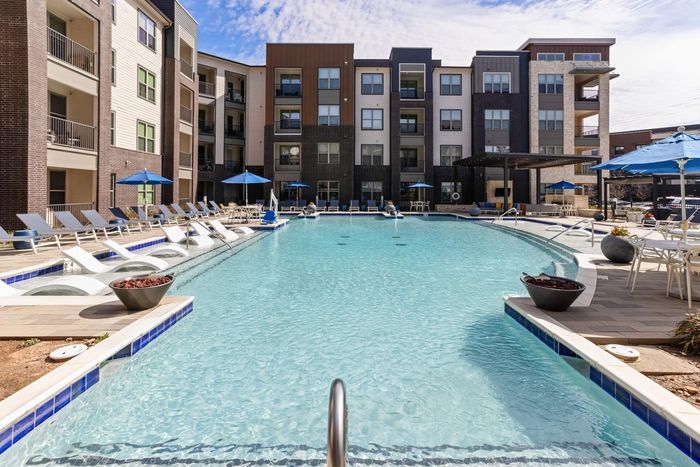 Luxurious apartment complex featuring a sparkling swimming pool with stylish lounge chairs and vibrant blue umbrellas, offering a perfect blend of comfort and elegance.