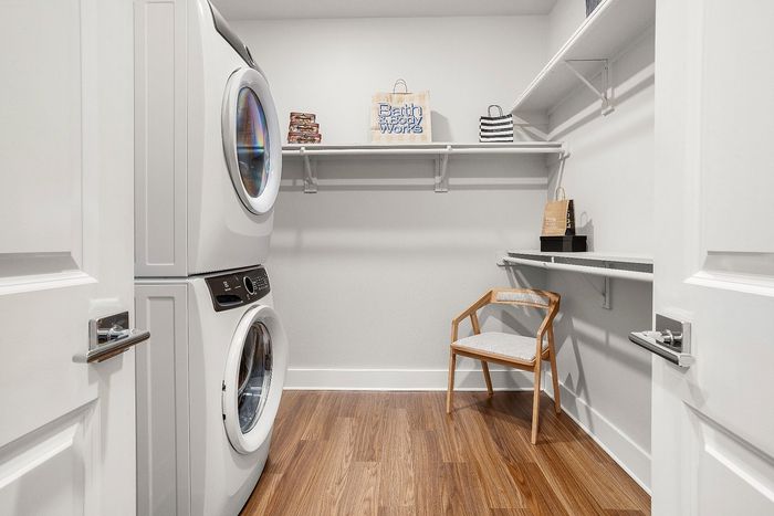 Modern laundry room with a stacked washer and dryer, elegant wood flooring, and ample storage shelves, featuring a chic wooden chair.