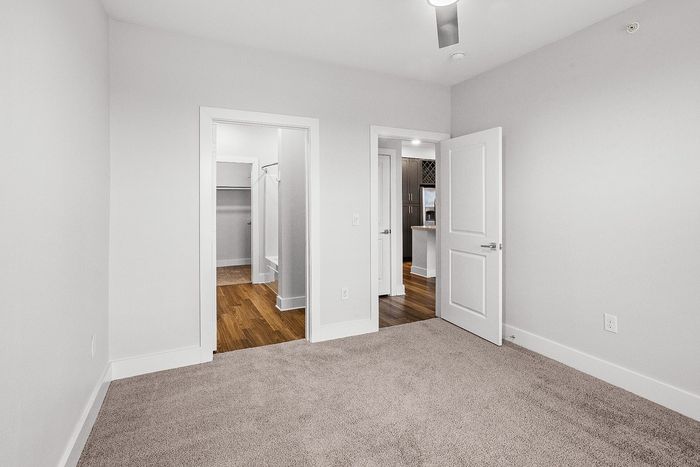 Bright and modern apartment room with plush carpet, sleek doors leading to a walk-in closet, and a view into a stylish kitchen with wood flooring.