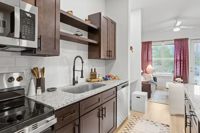 Modern kitchen with sleek granite countertops, espresso cabinets, and open shelving leads into a bright, cozy living area with large windows and stylish decor.