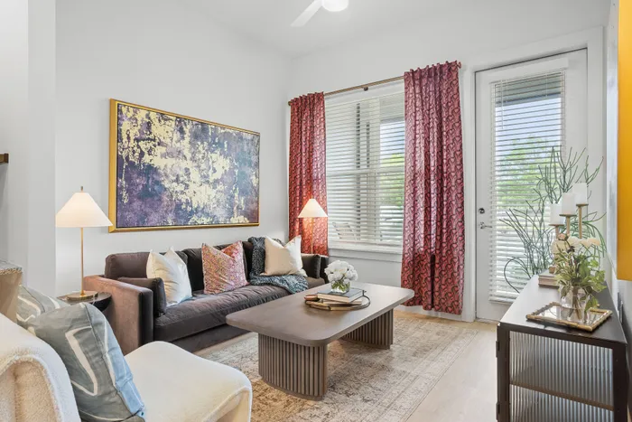 Inviting living room with modern decor, featuring a plush sofa, vibrant artwork, and natural light streaming through large windows with elegant red curtains.
