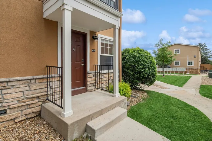Charming entrance to a modern townhouse with a private porch, lush green landscaping, and stone accents against a clear blue sky.