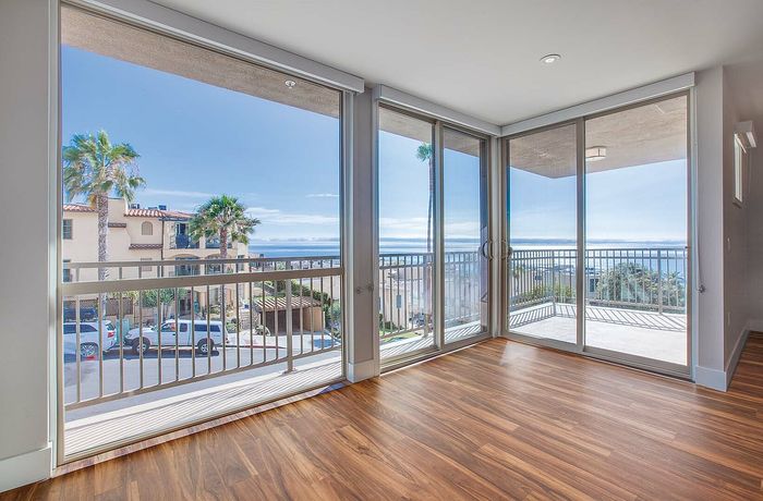 Bright apartment room with floor-to-ceiling windows offering stunning ocean views and hardwood floors, leading to a spacious balcony.