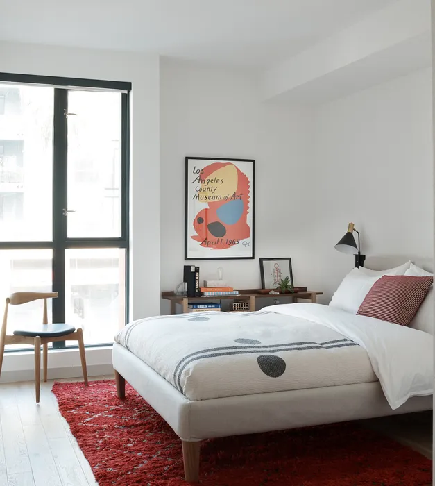 Modern bedroom with elegant decor, featuring a large window, cozy bed, stylish red rug, and chic art on the wall.