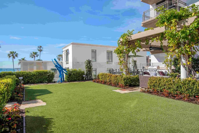 Luxurious apartment outdoor space featuring a lush green lawn, elegant pergola with climbing vines, stylish patio furniture, and vibrant flower beds against a backdrop of palm trees and ocean views.