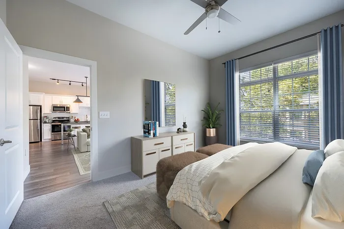 Bright and airy bedroom with modern decor, large window, and open view into a stylish kitchen with stainless steel appliances.