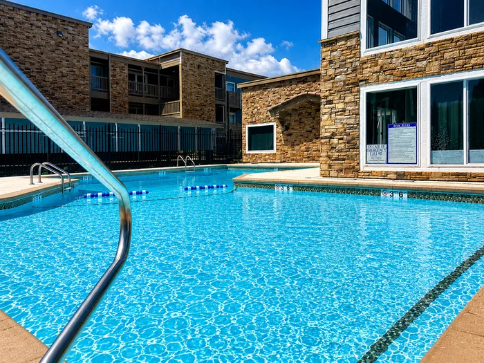 Luxurious apartment complex with sparkling blue swimming pool, surrounded by elegant stone architecture under a sunny sky.