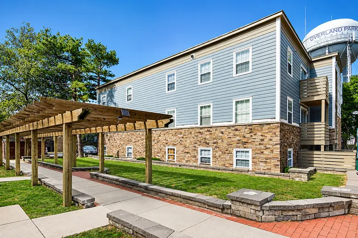 Charming residential building with rustic stone accents and a scenic wooden pergola walkway, nestled in lush greenery.