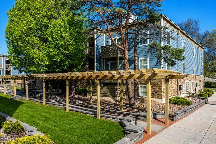 Charming apartment exterior with lush landscaping, elegant wooden pergola, and classic stone facade, promising a serene living environment.