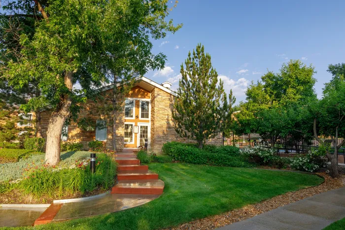 Charming stone-clad home with lush green landscaping, prominent trees, and a welcoming red entry path under a clear blue sky.