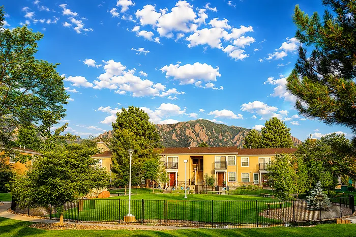 Charming apartment complex with lush green landscaping, vibrant trees, and stunning mountain backdrop under a clear blue sky.