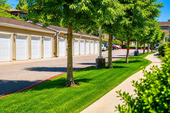Charming apartment complex featuring neat garages, a lush green lawn, and shaded walkways lined with vibrant trees.