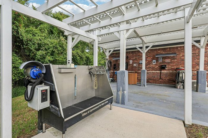 Modern outdoor pet wash center with stainless steel washing station under sleek pergola, surrounded by lush greenery and brick accents.
