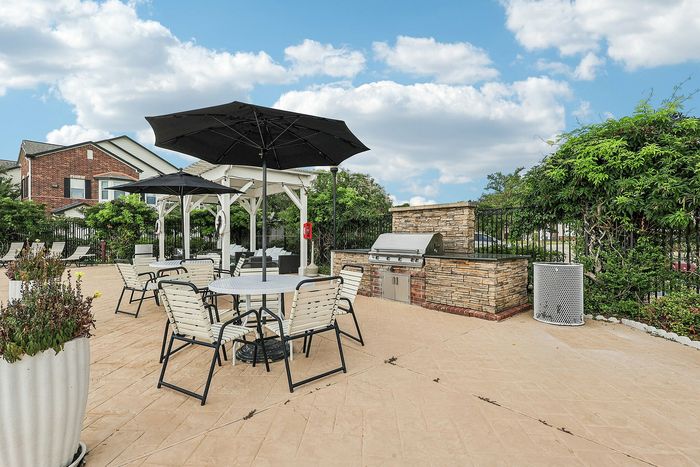 Charming outdoor patio with stylish seating, a barbecue grill area, and lush greenery under a bright blue sky.
