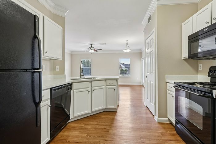 Bright and inviting kitchen with sleek black appliances, modern white cabinetry, and hardwood flooring, opening up to a spacious living area with elegant lighting.
