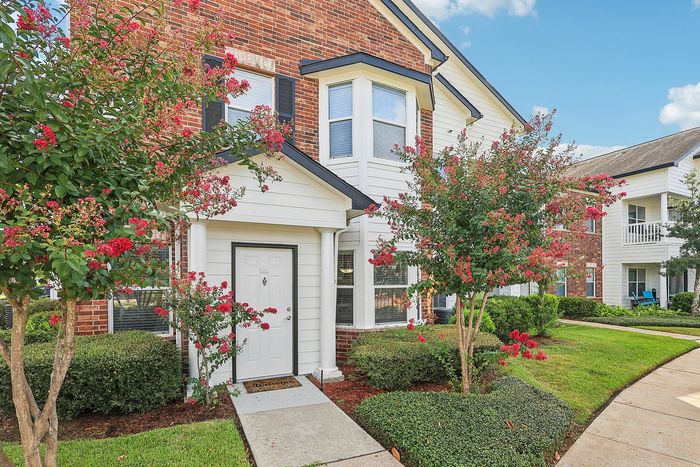 Charming townhouse with brick facade, lush landscaping, and vibrant red flowering trees, offering inviting curb appeal.
