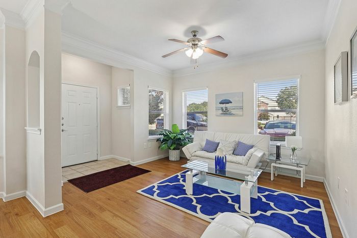 Inviting living area with modern decor featuring a sleek ceiling fan, plush white couch, vibrant blue rug, and large windows offering abundant natural light.