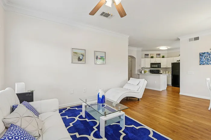 Bright living space featuring a cozy white sofa, stylish glass coffee table, and a vibrant blue rug, seamlessly flowing into a modern kitchen with sleek cabinetry.