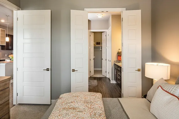 Inviting bedroom with elegant double doors leading to a chic walk-in closet and a modern en-suite bathroom, accented by warm lighting and stylish decor.