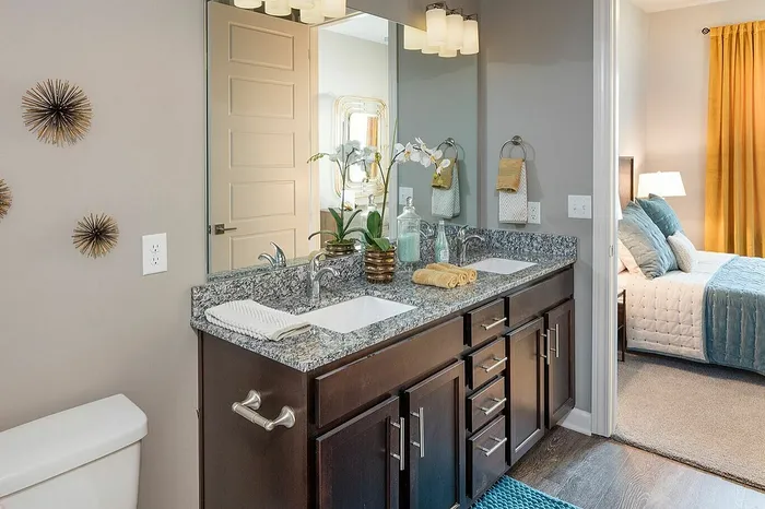 Elegant bathroom with dual sinks, granite countertop, and stylish decor, leading to a cozy bedroom with plush bedding and warm lighting.