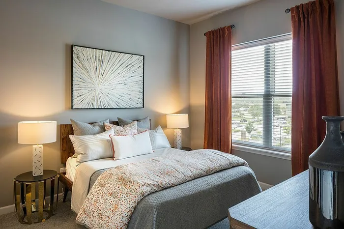 Inviting bedroom with modern decor, featuring a plush bed adorned with stylish cushions, a vibrant abstract painting, and warm natural lighting through elegant drapes.