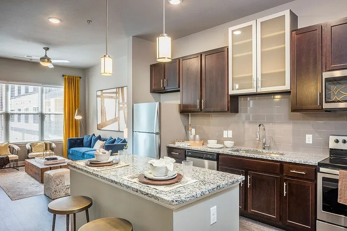 Modern apartment interior showcasing an elegant open-concept kitchen with granite countertops, adjacent to a cozy living area with a plush blue sofa and vibrant yellow curtains.
