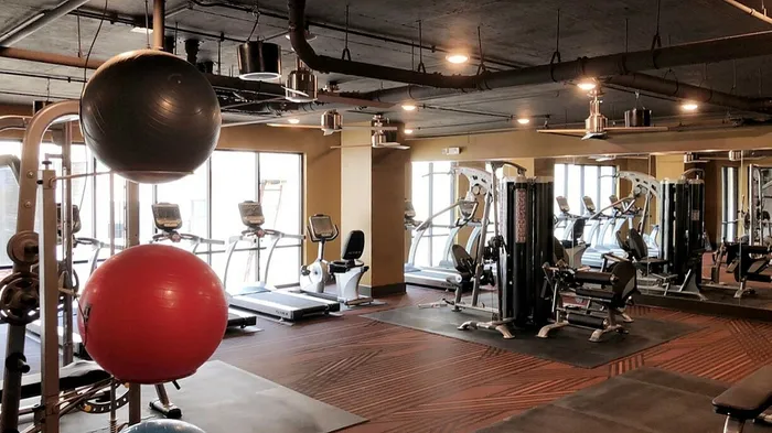 Modern fitness gym with state-of-the-art equipment including treadmills, exercise bikes, and weight machines, set in a spacious room with stylish flooring and large windows.